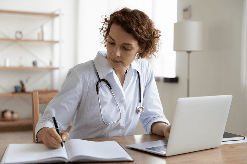 Serious female doctor using laptop and writing notes in medical journal sitting at desk. Young woman professional medic physician wearing white coat and stethoscope working on computer at workplace.