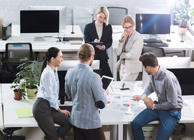 Five colleagues stood around table in bright office area with note pads, tablets and coffee using enterprise resource planning